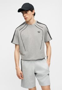 Νέος άνδρας που φοράει γκρι σόρτς και t-shirt Adidas με μαύρες ρίγες και μαύρο καπέλο, στέκεται με ένα χέρι στην τσέπη.