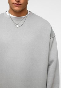 Grijze sweatshirt met een ronde hals, losse pasvorm en zichtbare textuur. Draag het over een wit shirt met twee zilveren kettingen als accenten.