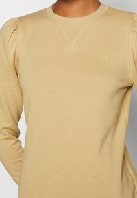 Svetr s dlouhými rukávy a světle béžovou barvou, který má nařeněné rukávy, žebrovaný výstřih a decentní logo. Vyroben z měkké, texturované tkaniny.