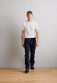 Witte korte mouwen t-shirt, marineblauwe broek en zwarte schoenen. Persoon draagt een marineblauwe pet met een logo. Achtergrond is lichtgekleurde en gestructureerde.