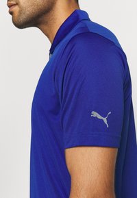 Polo bleu en tissu lisse, avec manches courtes, col, et logo Puma gris sur le bras gauche.