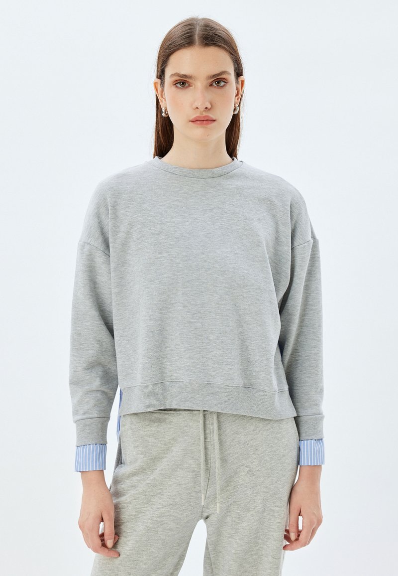 Sudadera gris con mangas largas, puños acanalados y un dobladillo corto, con acentos de rayas azules en los puños. Hecha de una tela suave.