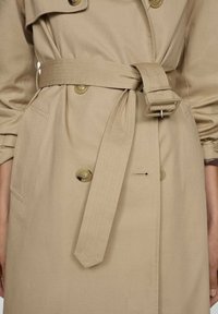 Beige trench coat with a belt, large buttons, two front pockets, and a tailored fit. Features a smooth texture and stitched details.