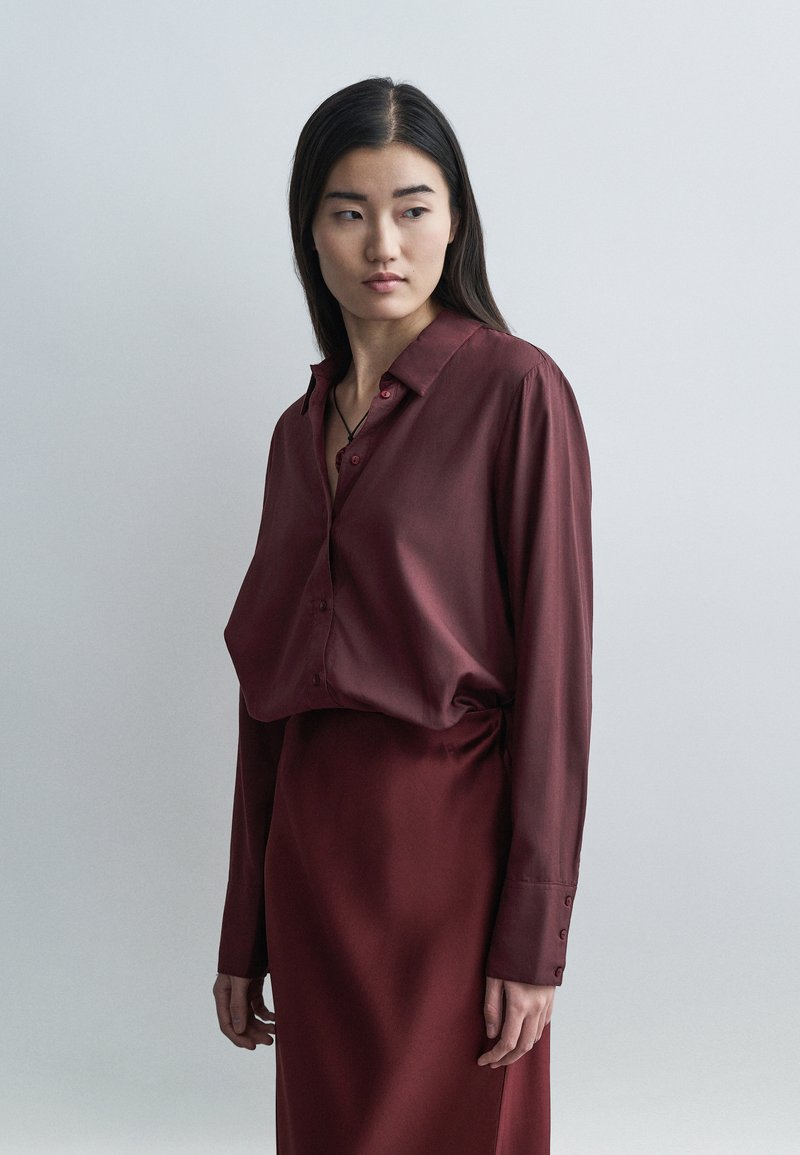Bordowa, koszula na długi rękaw z guzikami. Gładka tekstura z klasycznym kołnierzem, luźnym krojem i detalem z wszyciem po bokach. W zestawie z dopasowaną spódnicą.