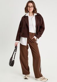 Giacca marrone doppiopetto, camicia bianca sotto, pantaloni larghi marrone a righe, e scarpe da ginnastica beige. Borsa nera con tracolla.