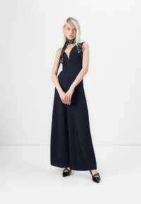 Navy jumpsuit met een diepe V-hals en wijde pijpen, gemaakt van een gladde stof. Aangevuld met een zwart-witte stippen sjaal om de hals.