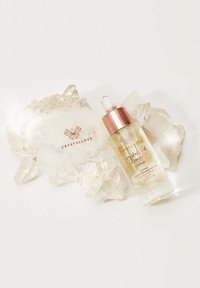 Bouteille en verre transparent avec un bouchon en or rose, contenant un sérum léger, à côté d'un masque facial en cristal lisse et transparent sur une surface blanche.
