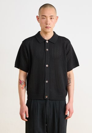 Cardigan corto nero a maniche corte lavorato a maglia con colletto, caratterizzato da un motivo aperto testurizzato e bottoni marroni lungo la parte anteriore.