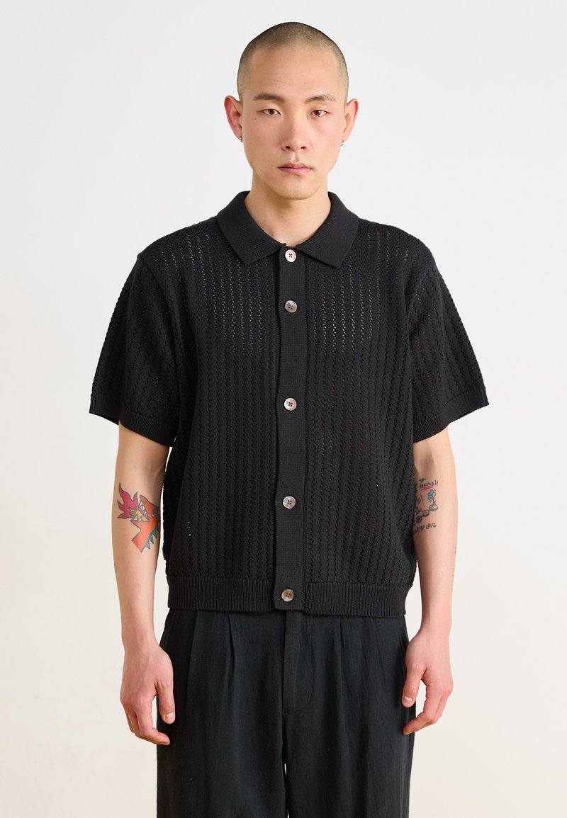 Black short-sleeve knitted cardigan with a collar, featuring a textured open pattern and brown buttons down the front.