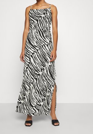 Frau trägt ein ärmelloses Maxikleid mit schwarz-weißem Zebramuster, einen seitlichen Schlitz und schwarze, offene Sandalen mit Absatz.