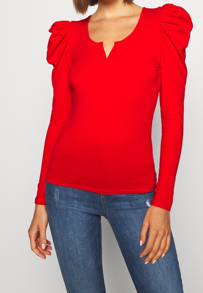 Maglietta rossa a maniche lunghe con spalle a palloncino e scollatura a V, realizzata in materiale elasticizzato. Abbinata a jeans blu con dettagli strappati.