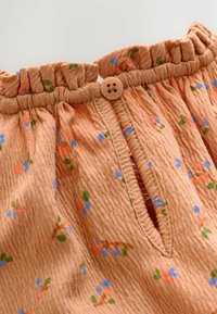 Tissu couleur pêche avec une texture froissée, présentant de petits motifs floraux bleus et orange. Comprend une fermeture à bouton et une taille froncée.