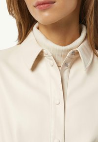 Ecrufarbene Kunstlederbluse mit einem strukturierten Kragen, Knopfverschluss und einem gerippten ecrufarbenen Rollkragenpullover darunter.