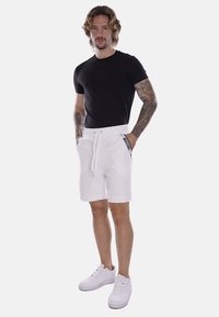 Pantaloni corti bianchi con una cintura a cordoncino e tasche laterali, con lettering del marchio nero. Abbinati a una maglietta nera aderente e sneakers bianche.