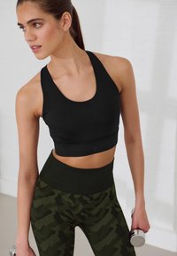 Top corto sportivo nero con scollatura rotonda, abbinato a leggings verde mimetico a vita alta. La modella tiene un manubrio.