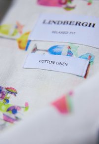 Farverig bomulds-linned stof med blomstermønstre. Mærkerne læser "Lindbergh," "Relaxed Fit," og "Cotton Linen." Blød tekstur, levende farver.