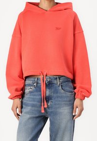 Koronkowy, skrócony hoodie w kolorze koralowym z troczkiem, ściągaczami i tonalnym logo. Zestawiony z niebieskimi dżinsami, o klasycznym pięciokieszeniowym fasonie.