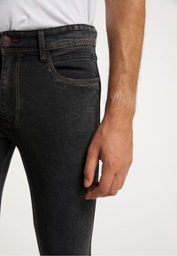 Jeans de mezclilla negra con corte ajustado, con costuras naranjas contrastantes, un bolsillo frontal y una textura suave. Mano visible al lado.