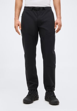 Άνδρας που φορά μαύρο παντελόνι υπαίθριου τύπου με ελαστικά μανσέτες και μαύρα ορειβατικά παπούτσια, στέκεται μπροστά σε ένα απλό λευκό φόντο.