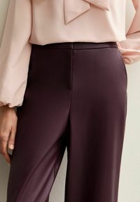 Burgundy, high-waisted pants made of smooth fabric, featuring a side pocket and tailored fit, paired with a light pink blouse with puff sleeves.