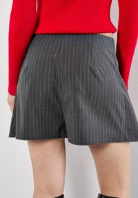 Donna che indossa una gonna grigia a righe sottili e un maglione rosso a coste, vista di schiena dalla vita alle ginocchia contro un muro semplice.