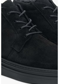 Czarne zamszowe buty z okrągłym noskiem, sznurowanym wzorem i teksturowaną czarną gumową podeszwą. Szczegóły obejmują subtelne akcenty przeszycia.