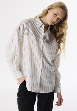 Camicia bianca a maniche lunghe con strisce verticali grigie e piccoli pois, con colletto e vestibilità morbida. Vengono indossati pantaloni neri.
