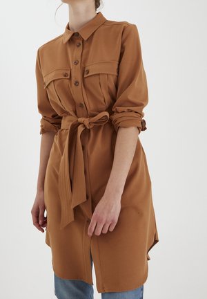 Vrouw draagt een lange bruine overhemdjurk met borstzakken en een gestrikte tailleband, gecombineerd met blauwe jeans. Mouwen opgerold.
