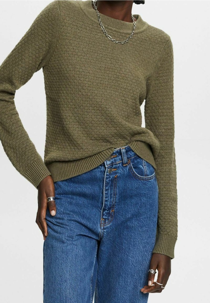 Personne portant un pull texturé vert olive, un jean taille haute bleu, un collier chaîne en argent et plusieurs bagues en argent aux deux mains.