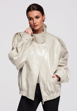 Kvinde iført stor, cremefarvet læderjakke med høj krave, knapper og elastisk kant, poserende med den ene hånd nær ansigtet og den anden i lommen.