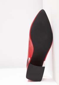 Chaussure rouge à bout pointu avec semelle noire et talon carré texturé, posée verticalement contre un mur d'angle blanc.