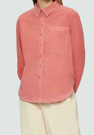 Kobieta ubrana w koralowo-różową sztruksową koszulę na guziki z kieszenią na piersi oraz kremowe, szerokie spodnie na nogawki na jednolitym tle.