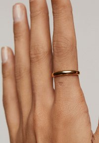 Anillo de metal dorado en una mano. El anillo tiene un diseño liso y redondeado con una superficie reflectante, en un fondo claro.