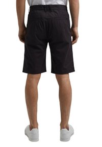 Homme portant un short noir au niveau des genoux, des baskets blanches et une chemise claire, vu de dos sur fond blanc.