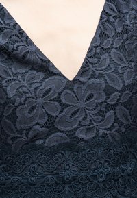 Κοντινή λήψη ενός navy μπλε δαντελένιου φορέματος με λουλουδένια σχέδια και V-σχήμα λαιμόκοψης που αποκαλύπτει ανοιχτόχρωμο δέρμα πάνω από το στήθος.