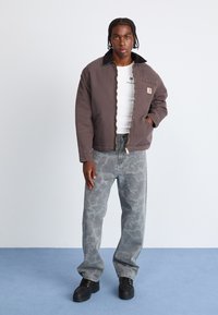 Brun kanvasjakke med svart krage, hvit T-skjorte under, grå mønstrede jeans og svarte snøreluer. Modellen står med fronten vendt mot kamera.