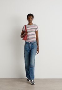 T-shirt rayé avec des lignes horizontales rouges, bleues et blanches, associé à un jean bleu ample. Sac à main rouge, baskets noir et blanc. Fond simple.
