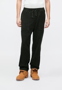 Pantaloni din catifea côtelată verde închis, cu talie elastică, buzunare frontale și design cu picior drept, asortați cu bocanci maro deschis cu șireturi.