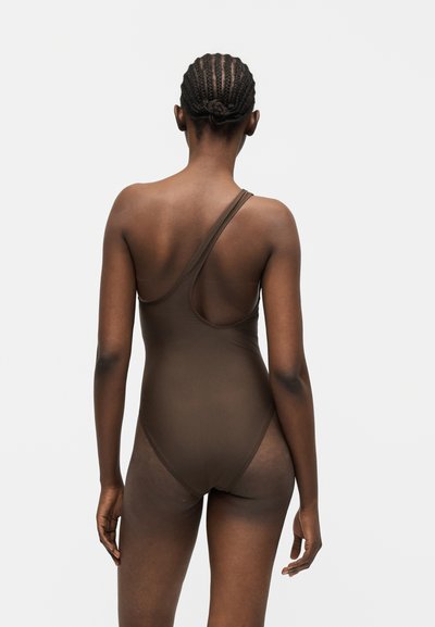 Γυναίκα με πλεξούδες που φοράει καφέ ολόσωμο μαγιό με ένα ώμο, στέκεται γυρισμένη με την πλάτη της προς ένα απλό ανοιχτό φόντο.