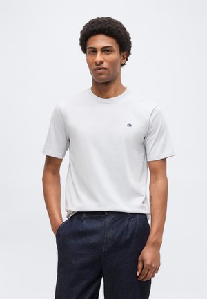 Homme aux cheveux courts et bouclés portant un t-shirt uni gris clair et un pantalon bleu foncé, debout avec une main dans la poche.