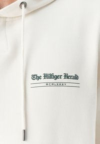 Biela mikina s jemnou textúrou, na prednej strane s potlačeným zeleným textom: "The Hilfiger Herald" a "MCMLXXXV", doplnená kovovou šnúrkou.