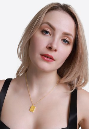 Gouden ketting met een rechthoekige hanger met een hoekig ontwerp, hangend aan een delicate ketting, gedragen over een zwarte stoftop.