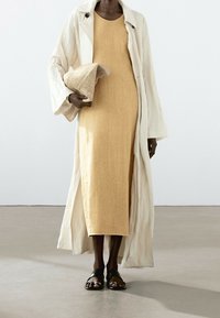 Leinen-gelbes Kleid mit einem langen, weißen Trenchcoat; hält eine strukturierte beiger Clutch und trägt schwarze Sandalen.