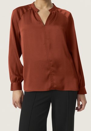 Rostfarbene Bluse mit Mock-Neck-Kragen, langen puffärmeln und subtilen Texturmusterungen. Kombiniert mit schwarzen Hosen für einen Kontrast.