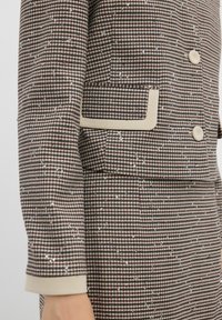 Brązowa i beżowa marynarka w kratę z cekinami, pojedynczą kieszenią z beżowym wykończeniem i beżowymi guzikami na przodzie.