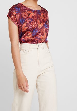 Vrouw draagt een satijnen top met korte mouwen in rood, oranje en blauw met bladpatronen, gecombineerd met een high-waisted off-white broek.