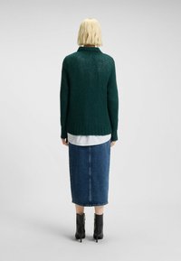 Maglione a collo alto di colore verde scuro, abbinato a una camicia bianca e una gonna di jeans, completato da stivali alla caviglia neri.
