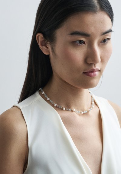 Νεαρή γυναίκα με μακριά σκούρα μαλλιά, φορεί λευκή αμάνικη μπλούζα και ένα κολιέ από μαργαριτάρια και κρύσταλλα, κοιτάζει προς τα κάτω με ουδέτερη έκφραση.