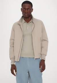 Hellbeige Bomberjacke mit hohem Kragen, Reißverschluss vorne und gerippten Bündchen, getragen über einem hellgrauen Poloshirt und blauen Cordhosen.