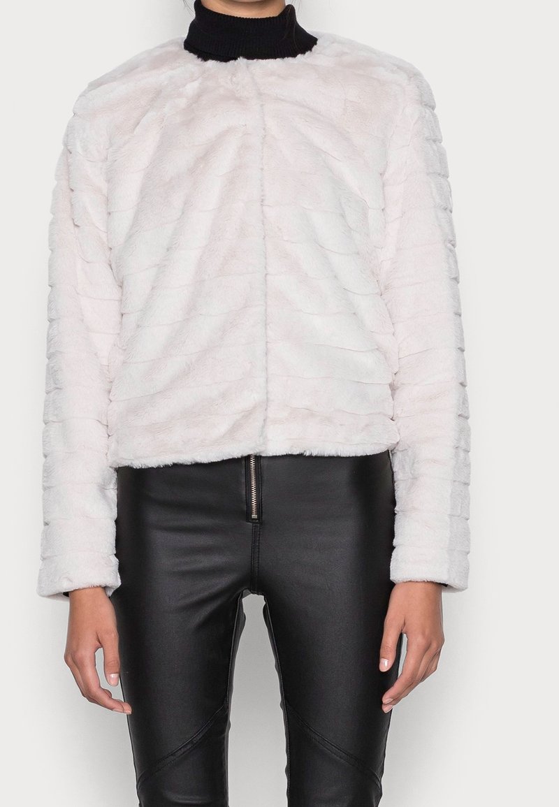 Femme portant une veste en fausse fourrure blanche texturée sur un col roulé noir associée à un pantalon en cuir noir taille haute avec fermeture éclair à l'avant.